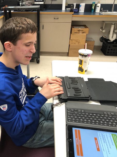 student sits at a table and reads a passage on his Braille Note Touch
